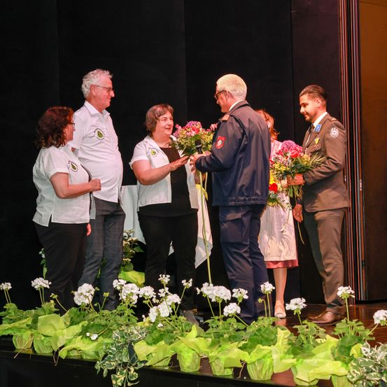 Eine Gruppe von Menschen, darunter zwei Frauen und zwei Männer in Uniformen, steht auf der Bühne, hält Blumen und lächelt. Die Bühne ist mit weißen Blumen und grünen Blättern geschmückt.