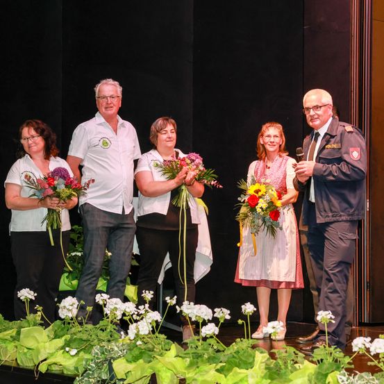 Fünf Personen auf der Bühne, alle lächelnd, Blumen haltend. Ein Mann im Anzug steht rechts und hält ein Mikrofon. Die Frauen tragen Kleider und die Männer weiße Hemden. Der Hintergrund ist schwarz.