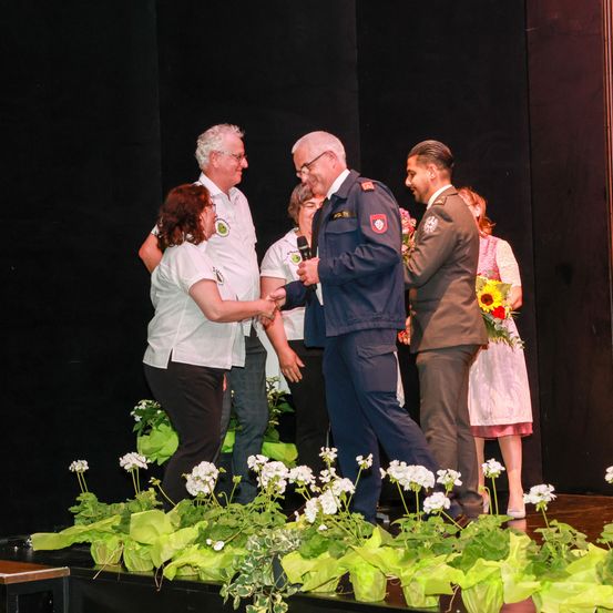 Eine Gruppe von Menschen steht auf einer Bühne mit weißen Blumen. Ein Mann in Uniform hält ein Mikrofon. Ein anderer Mann hält einen Blumenstrauß. Eine Frau schüttelt die Hand eines Mannes in Uniform.
