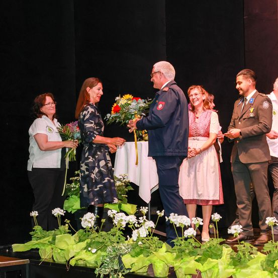Ein Mann in Uniform überreicht einer Frau auf der Bühne Blumen, in der Nähe stehen andere Frauen und ein Soldat. Die Bühne ist mit Pflanzen und Blumen dekoriert.