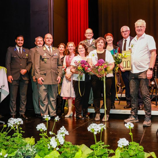 Eine Gruppe von Menschen steht auf der Bühne, einige halten Blumen und andere Auszeichnungen. Sie sind in verschiedenen Uniformen gekleidet und scheinen für ein Foto zu posieren.
