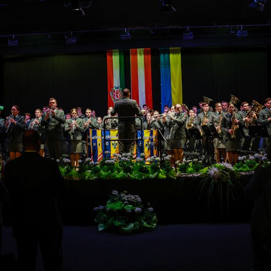 Ein Militärorchester tritt bei Nacht auf der Bühne auf, geschmückt mit bunten Fahnen und Blumen. Die Mitglieder in Uniform spielen Trompeten, Flöten und Posaunen.