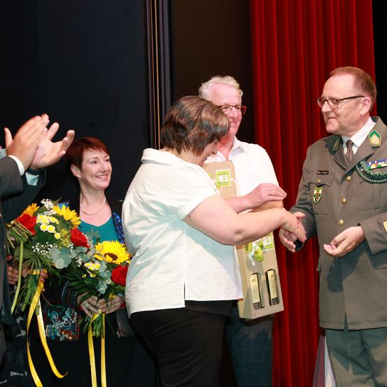 Eine Frau in einem weißen Hemd erhält ein Geschenk von einem Mann in Uniform. Sie sind auf einer Bühne mit roten Vorhängen, und eine andere Person klatscht.
