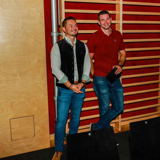 Zwei Männer stehen nebeneinander in einem Raum. Der Mann auf der linken Seite trägt eine Weste und Jeans, während der Mann auf der rechten Seite ein rotes Poloshirt trägt und eine schwarze Mappe in der Hand hält.
