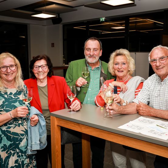 Eine Gruppe älterer Menschen lächelt und posiert für ein Foto mit Wein glasses in der Hand. Ein Tisch vor ihnen hat Wein glasses, eine Flasche und Servietten.
