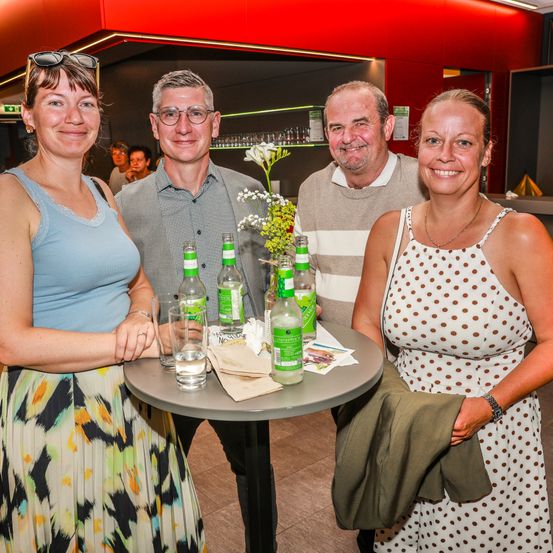 Vier Menschen lächeln und posieren für ein Foto. Der Mann links trägt eine Brille und ein graues Hemd. Die Frau neben ihm trägt ein ärmelloses Oberteil. Der Mann in der Mitte hat ein gestreiftes Hemd. Die Frau rechts trägt ein Kleid mit Punkten. Sie stehen vor einem Tisch mit grünen Flaschen und Blumen.