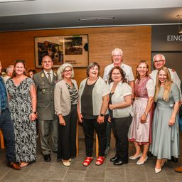 Eine Gruppe von Menschen, einschließlich Militäroffiziere, steht in einem Raum mit einer Holzwand. Sie alle lächeln und schauen zur Kamera. Der Raum hat einen gefliesten Boden und eine Decke mit Lampen.