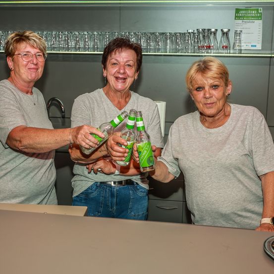 Drei ältere Frauen lächeln in einer Küche und halten Flaschen eines Getränks. Hinter ihnen befinden sich Regale mit verschiedenen Gläsern und einer Preisliste.