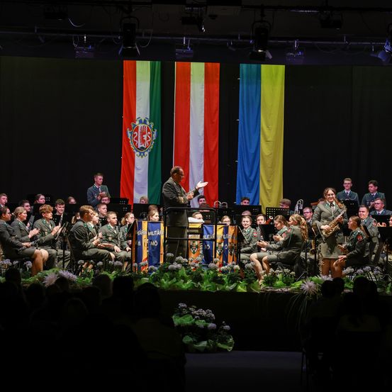 Ein Militärorchester auf der Bühne, mit einem Dirigenten, Musikern und Publikum. Flaggen im Hintergrund.