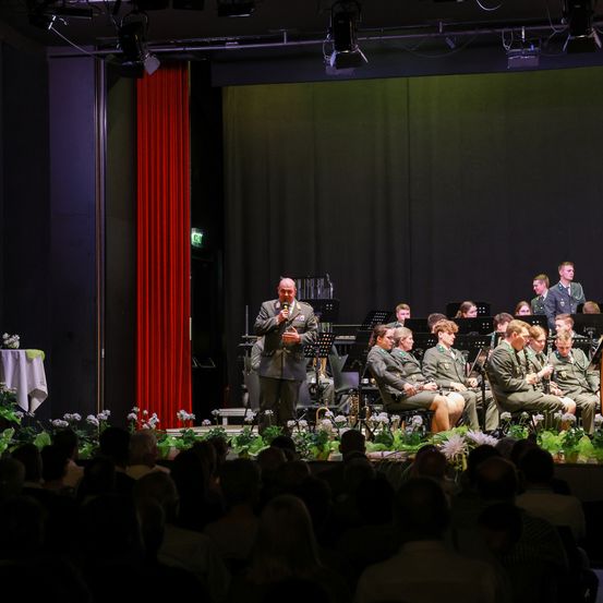 Ein Militärorchester spielt auf einer Bühne mit roten Vorhängen, Blumen und einem Publikum, wobei ein Mann in ein Mikrofon spricht.