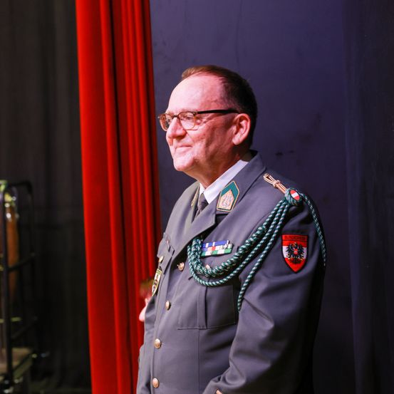 Ein älterer Mann in Uniform mit Brille steht vor einem roten Vorhang. Er trägt Orden auf der Brust und eine grüne Schärpe.