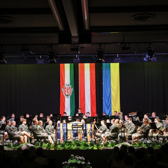 Ein Militärorchester in Uniform tritt auf der Bühne auf, mit Flaggen aus Italien und der Ukraine im Hintergrund. Das Publikum sitzt vor ihnen.