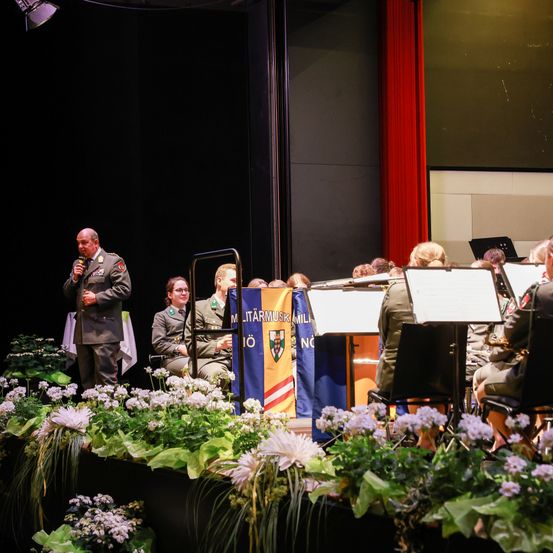 Ein Militärorchester spielt auf der Bühne. Ein Mann in Uniform spricht in ein Mikrofon. Die Band sitzt und spielt Instrumente. Ein Fahnenmast steht hinter ihnen.