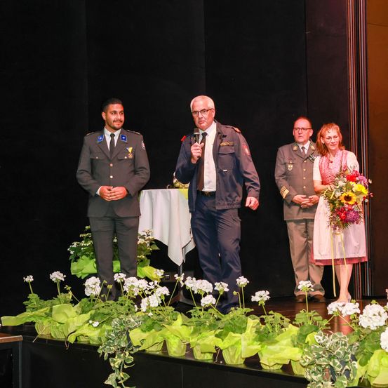 Ein Mann auf der Bühne hält ein Mikrofon in der Hand und spricht zu drei anderen in Uniform, einer hält einen Blumenstrauß. Dahinter befindet sich eine Kulisse in dunklen Farben und ein Tisch mit einer weißen Tischdecke.