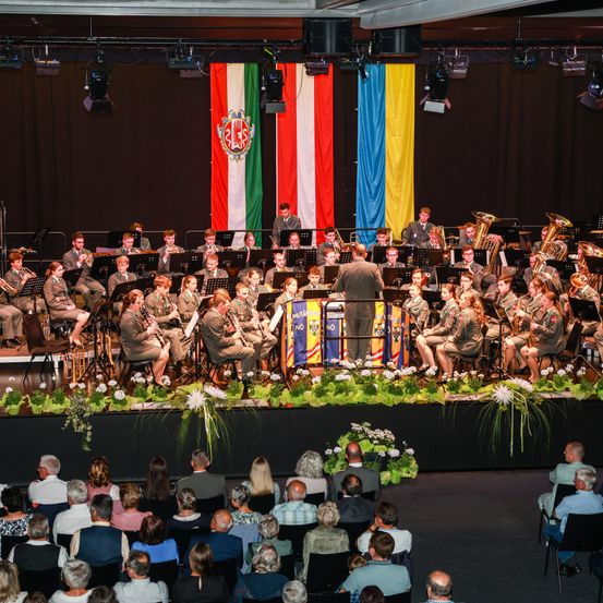 Ein Militärorchester spielt auf der Bühne mit verschiedenen Instrumenten unter Flaggen. Das Publikum sitzt vor der Bühne und schaut zu.