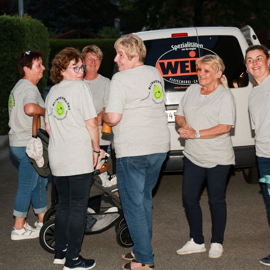 Eine Gruppe von Frauen in passenden T-Shirts steht vor einem Van mit dem Schriftzug 'Spezialist für Blindenmarkt.' Sie scheinen miteinander zu sprechen.