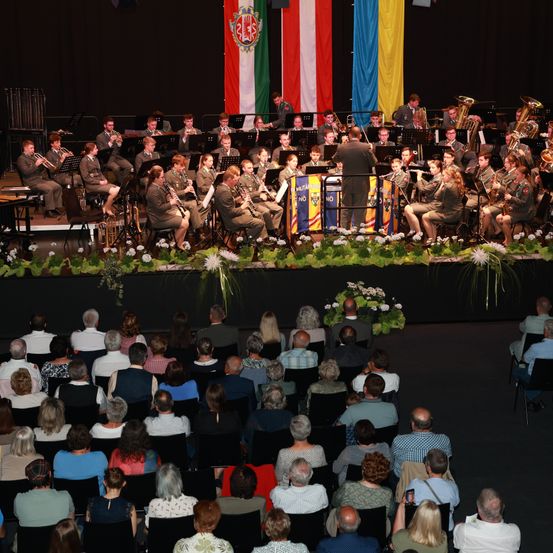 Ein Militärorchester spielt auf der Bühne mit verschiedenen Instrumenten wie Trompeten und Tuben. Fahnen von Ungarn und einem anderen Land schmücken den Hintergrund. Ein Publikum sieht vom Sitzbereich aus zu.