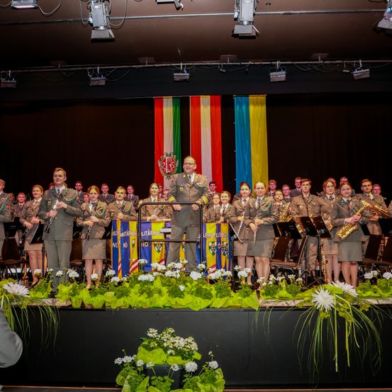 Ein Militärorchester steht auf der Bühne und spielt verschiedene Instrumente. Ein Mann in Uniform steht in der Mitte und spricht das Publikum an. Banner in den Farben Rot, Weiß und Blau hängen im Hintergrund.