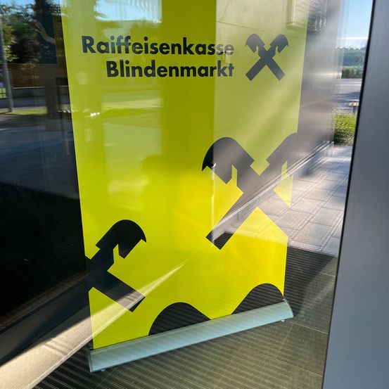 Ein gelbes Banner mit schwarzem Text und Symbolen hängt an einer Glastür. Es steht Raiffeisenkasse Blindenmarkt mit drei schwarzen gekreuzten Hämmern.