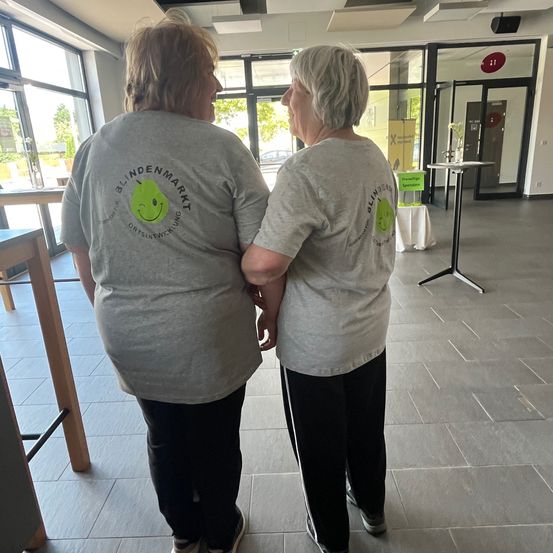 Zwei Frauen mit grauen T-Shirts und schwarzen Hosen stehen und schauen in einen offenen Raum.