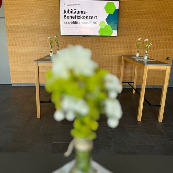Ein Raum mit Holzwänden und -böden. Zwei Tische haben Vasen mit weißen Blumen. Ein Monitor zeigt eine Nachricht über ein Jubiläums-Benefizkonzert mit Militärmusik.