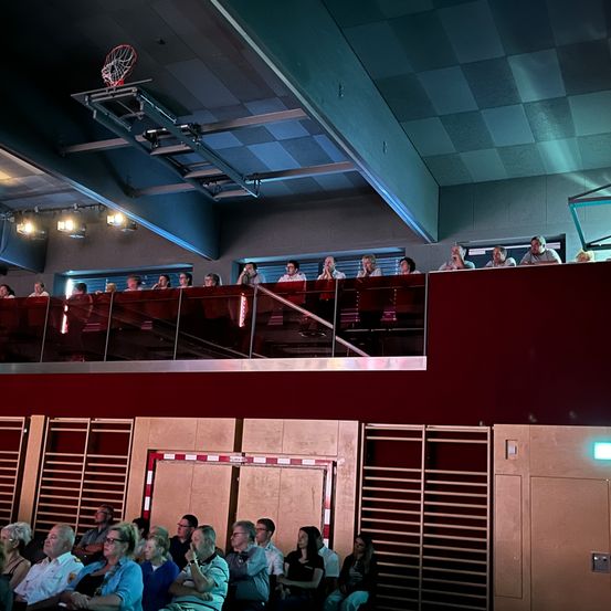 Eine Indoor-Basketballhalle mit einigen Leuten auf der Galerie. Einige Menschen sitzen im Erdgeschoss. Die Decke ist weiß und blau. Ein Basketballkorb ist an der Decke befestigt.