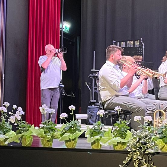 Ein Mann fotografiert eine Band auf der Bühne. Die Musiker spielen Trompeten und Flöten. Sie sind von Blumentöpfen umgeben.