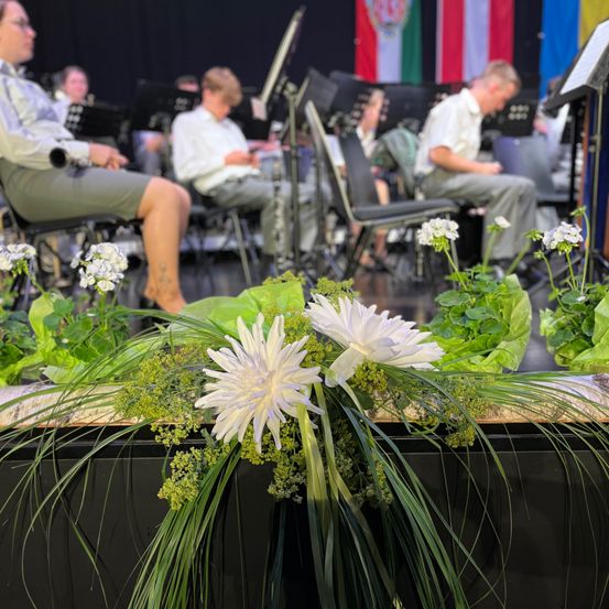 Ein Orchester spielt auf einer Bühne mit Blumen und Grünpflanzen davor. Die Musiker sitzen, tragen weiße Hemden und graue Hosen, und Flaggen sind im Hintergrund sichtbar.