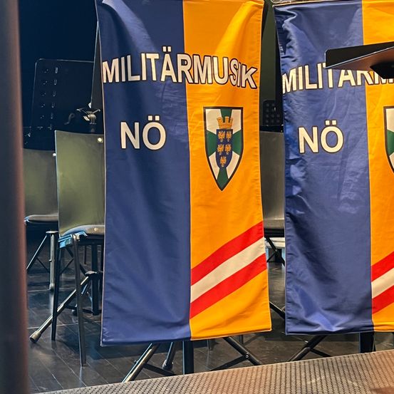 Zwei blaue und gelbe Banner mit weißem Text und Logo sind auf einer Bühne ausgestellt, mit Stühlen und einem Podium im Hintergrund.