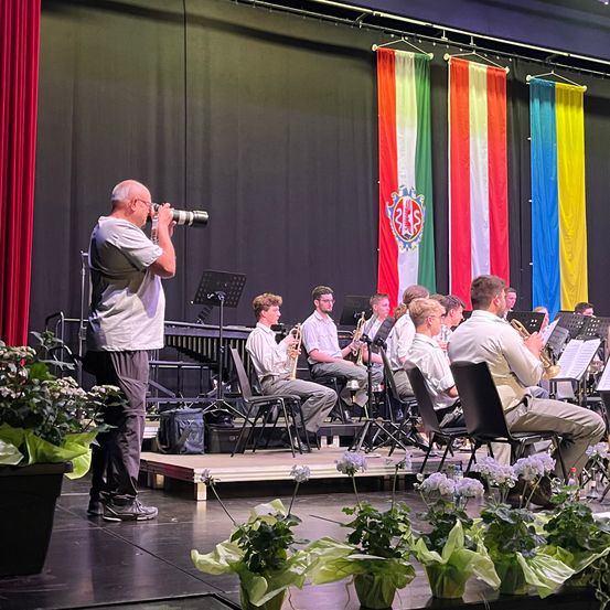 Ein Mann fotografiert die Darbietung einer Band auf der Bühne, umgeben von Flaggen und Topfpflanzen. Die Musiker spielen Trompeten und Posaunen und sitzen auf Stühlen.