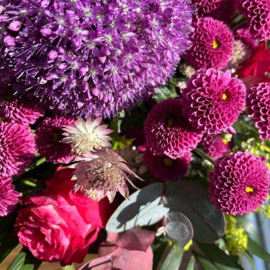 Eine Nahaufnahme einer Blumenarrangement mit verschiedenen Blumen, darunter rosa und lila Blüten, eine rote Rose und grüne Blätter. Das Arrangement ist lebendig und farbenfroh.