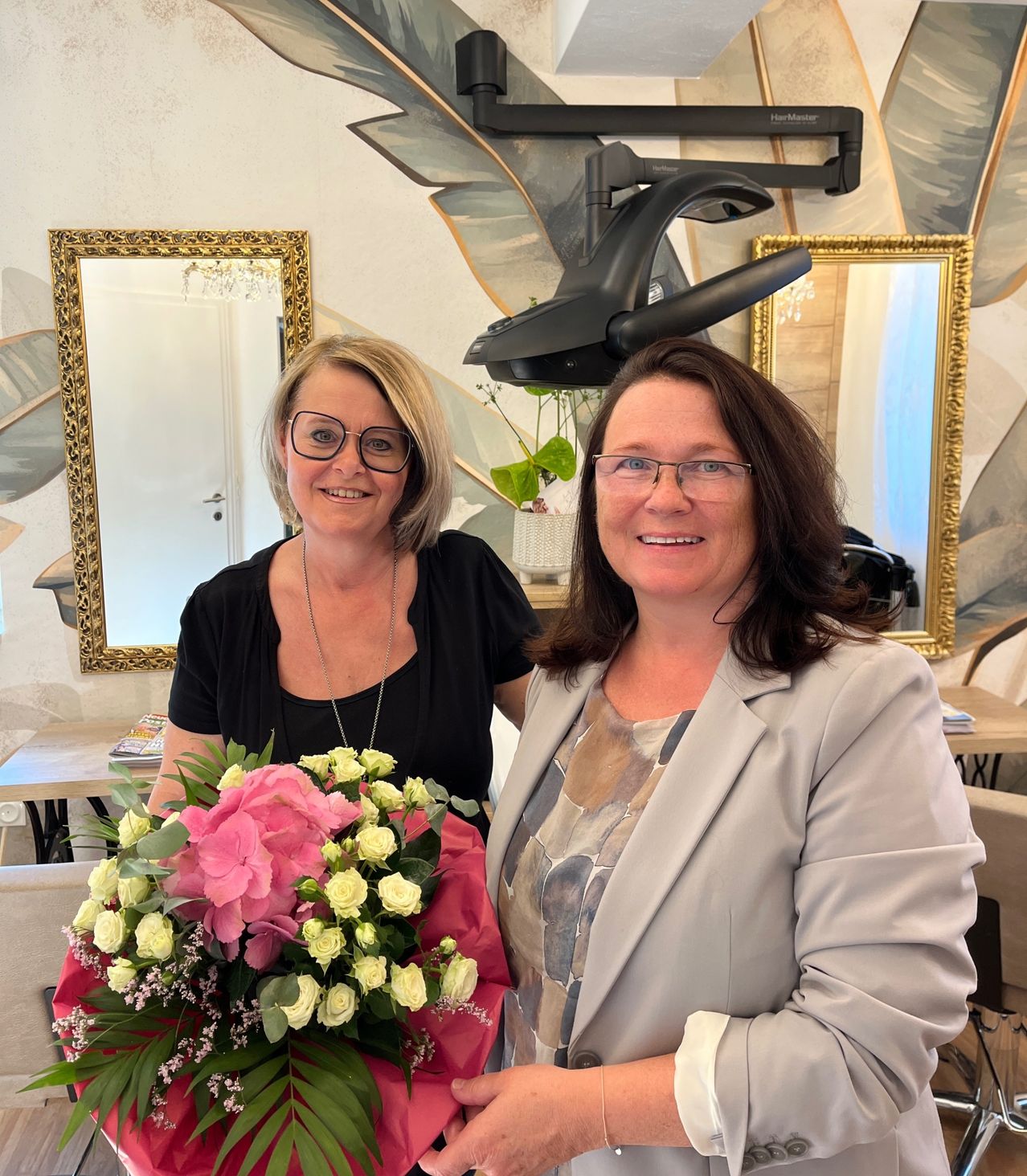 Zwei Frauen stehen nebeneinander in einem Raum mit einer dekorativen Wand. Die Frau auf der linken Seite hält einen Blumenstrauß. Hinter ihnen befindet sich ein Spiegel und eine Pflanze in einer weißen Vase.