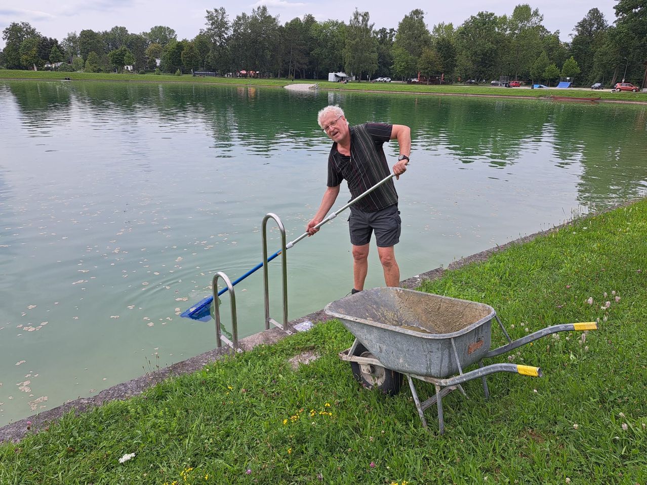 Ein älterer Mann hält eine Harke am Ufer eines Teichs, ein Schubkarren steht in der Nähe, möglicherweise zur Reinigung des Wasserrands.