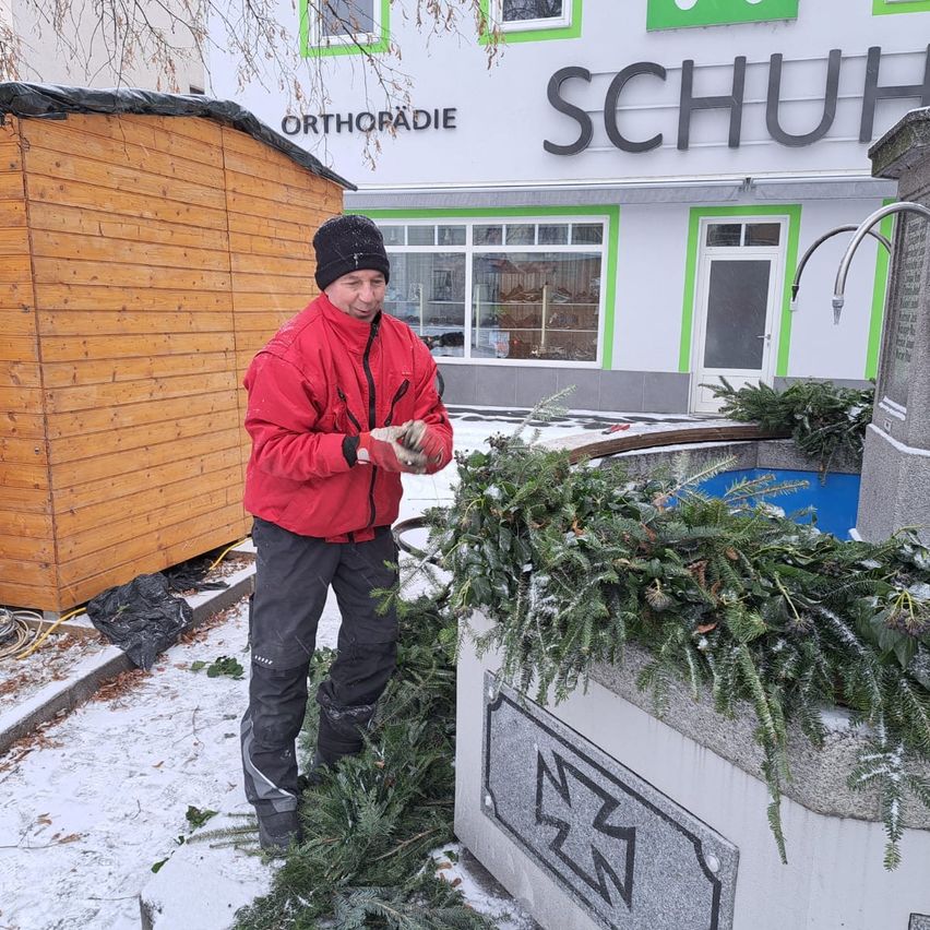 Ein Mann in einer roten Jacke schmückt einen Brunnen mit Tannenzweigen in einem verschneiten Bereich vor einem Gebäude mit dem Schild 'SCHUH ORTHOPÄDIE'.