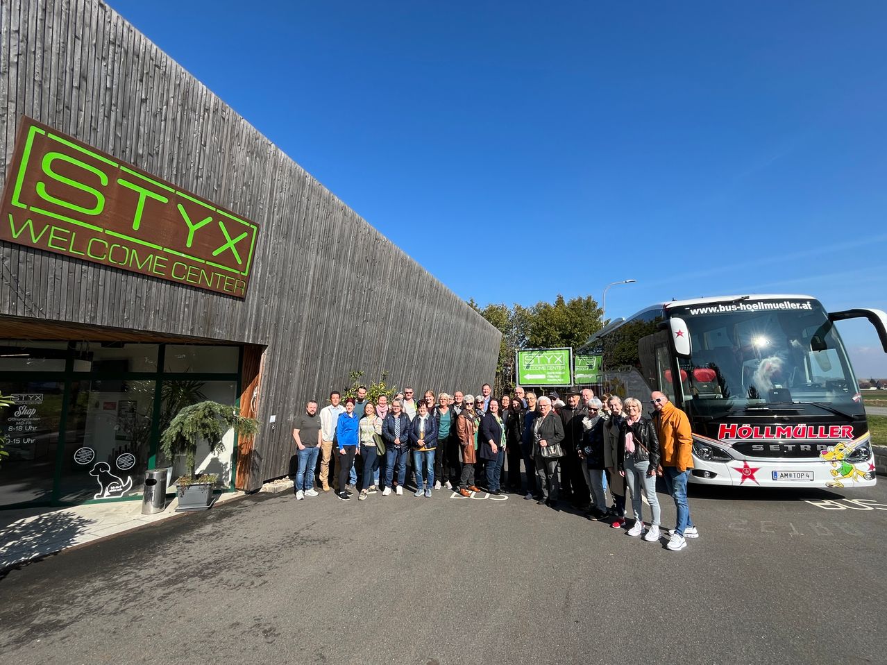 Eine Gruppe von Menschen steht vor einem Gebäude mit der Aufschrift 'TYX HOME CENTER', mit einem Bus mit der Aufschrift 'HOLL' dahinter.
