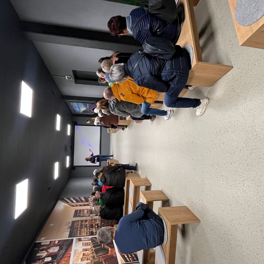 Eine Aufsicht von Menschen, die auf Bänken in einem Raum sitzen. Eine Person steht vor einer Projektionsleinwand. Die Wände sind schwarz mit Bildern.