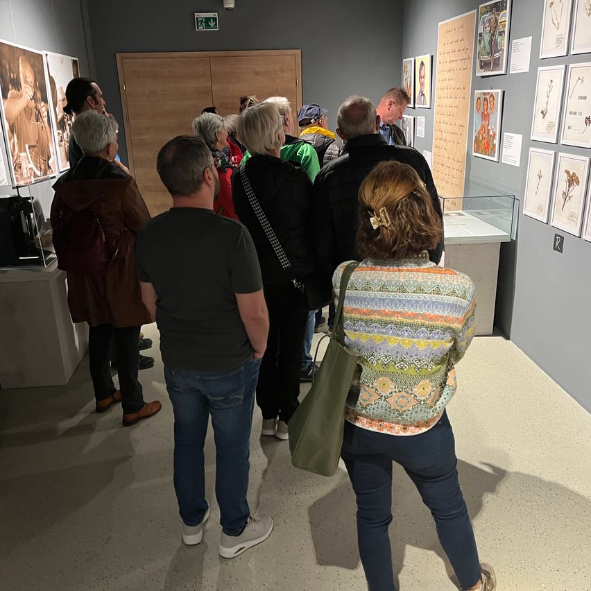 Eine Gruppe von Menschen steht in einer Kunstgalerie und betrachtet gerahmte Gemälde und Drucke an der Wand. Einige tragen Jacken und Turnschuhe. Ein Glaskasten hält Objekte auf der rechten Seite.