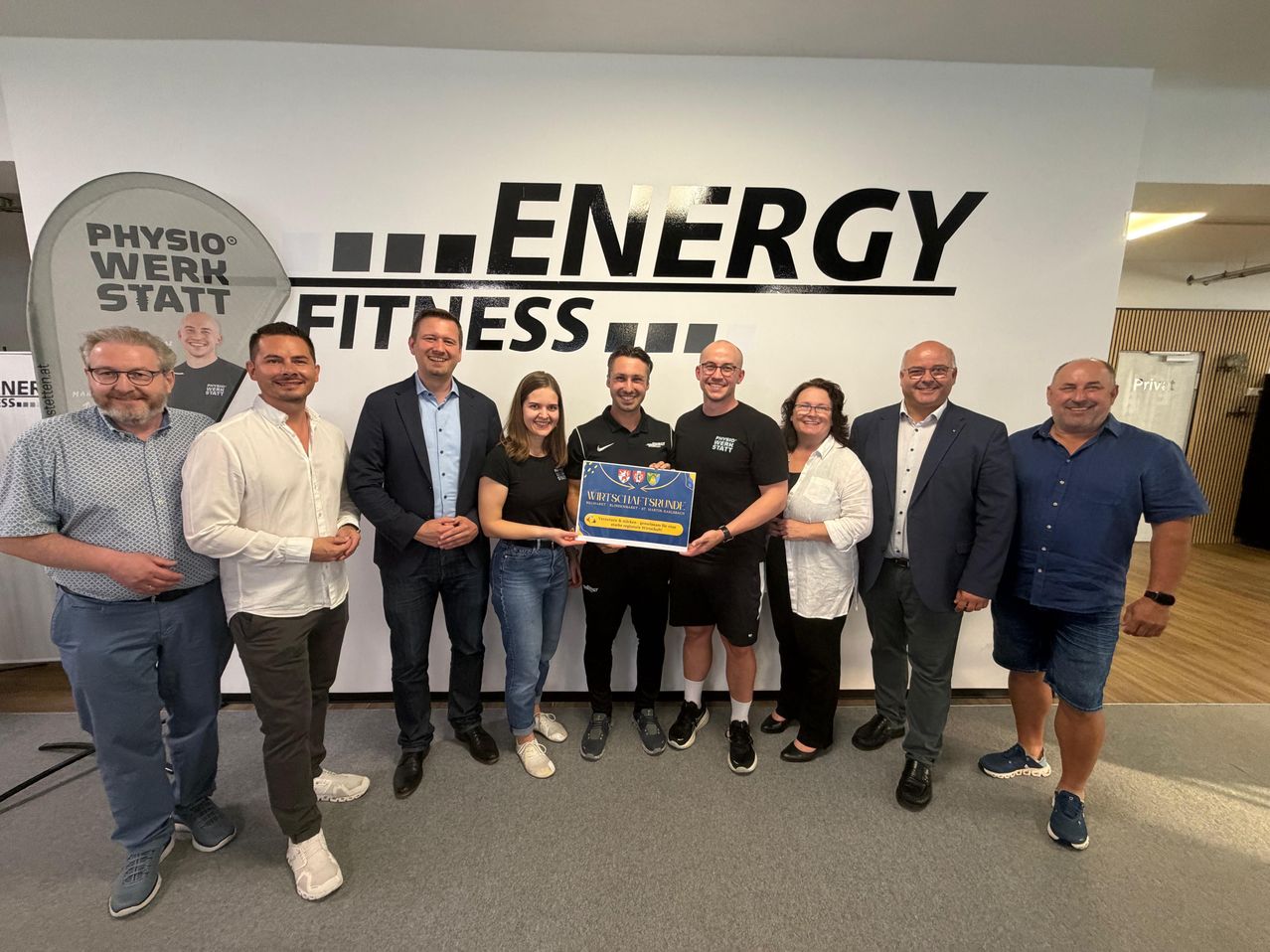 A group of people pose for a photo in front of a wall with the word 'Energy Fitness' on it. They are smiling and holding a certificate.