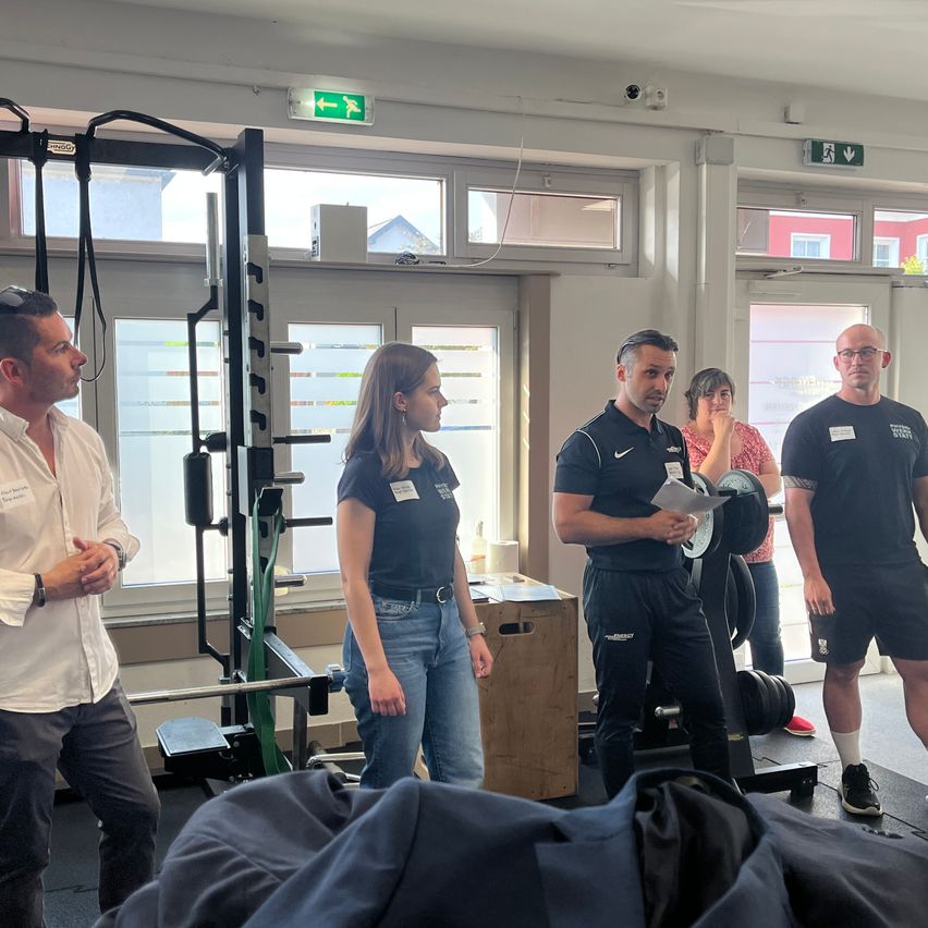 Five people stand in a gym, with two men holding papers and the others listening. A woman in a white shirt faces the man holding papers. A barbell and weights are on the ground.