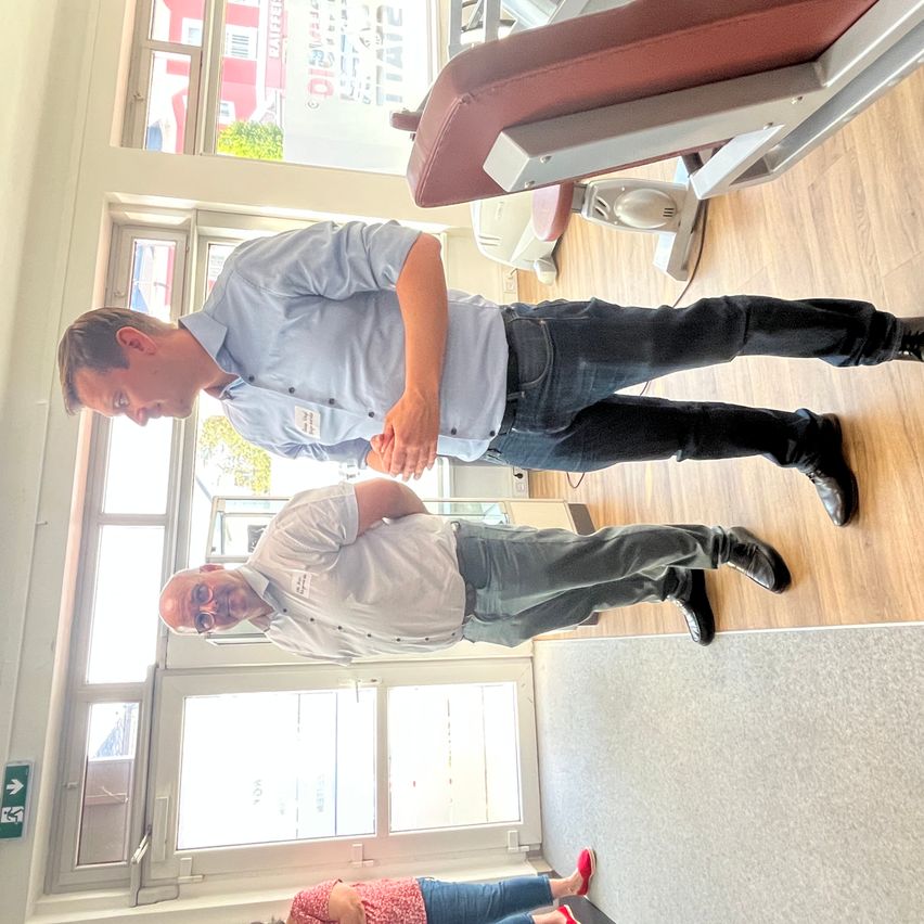 Two men are standing in a gym. One man is wearing a blue shirt, and the other is wearing a white shirt. They are looking at something on the ground. A woman is sitting on a chair. There are gym equipment in the background.