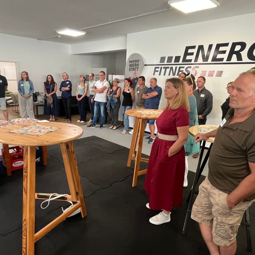 In a gym room, several people stand around a table playing a board game. The room has a black floor mat and a wall with the words 'ENERGY FITNESS'.