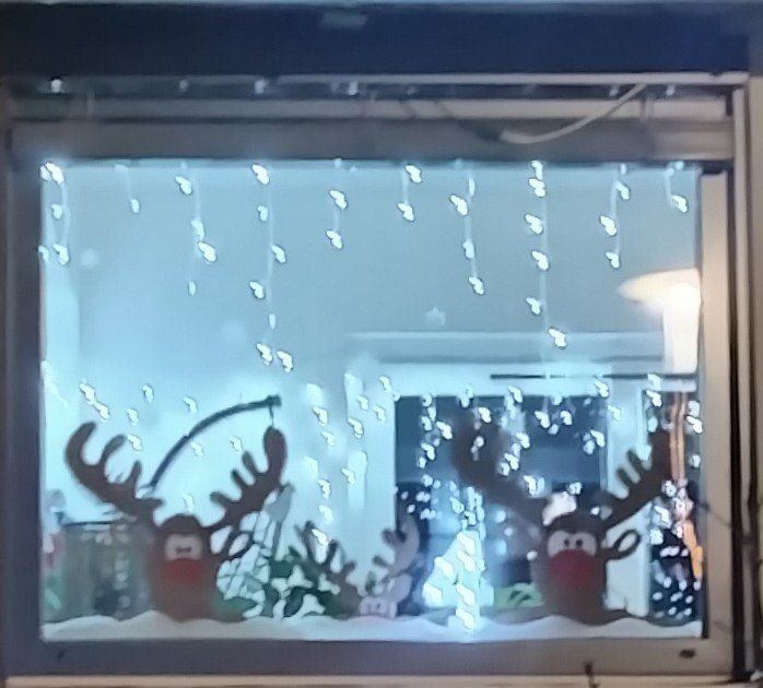 Ein Fenster zeigt eine festliche Szene mit zwei Rentieren unter Schneefall. Lichter und Dekorationen sind durch das Glas sichtbar.