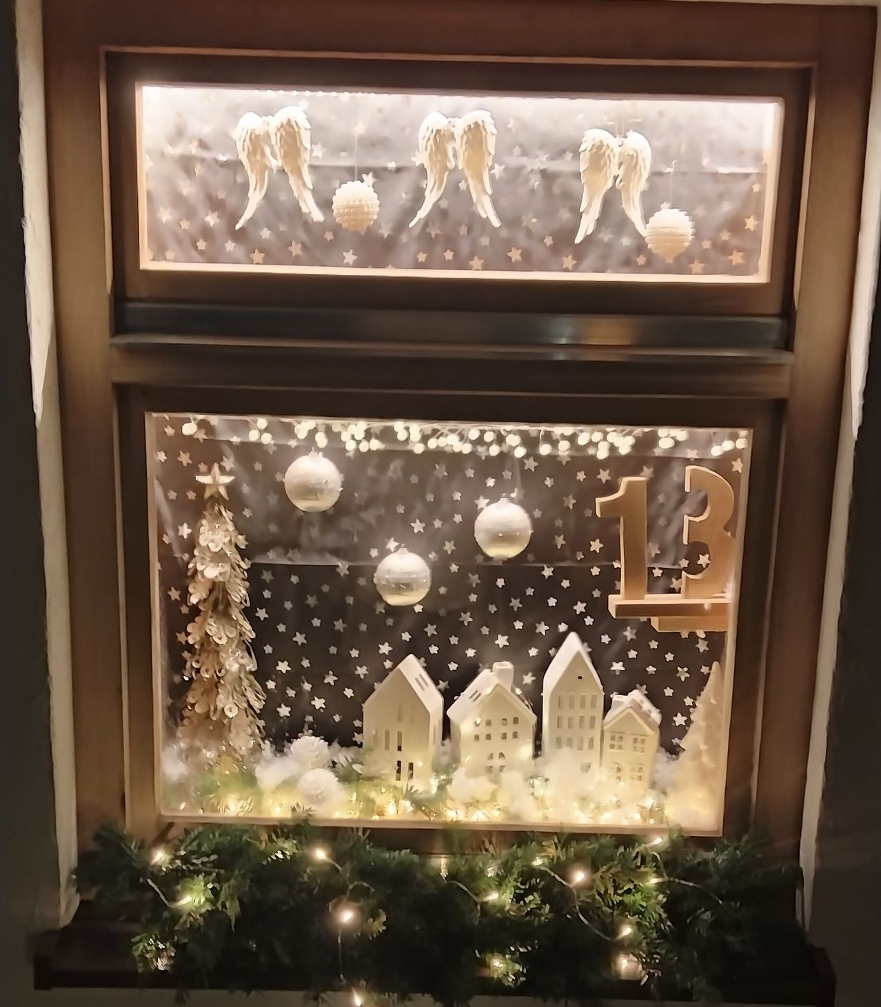 Ein Fensterdisplay mit einer verschneiten Szene, bestehend aus einem Weihnachtsbaum, Häusern und hängenden Ornamenten, beleuchtet von Lichtern. Der obere Teil hat Engelsflügel und Sterne. Das Datum '13' ist prominent angezeigt.