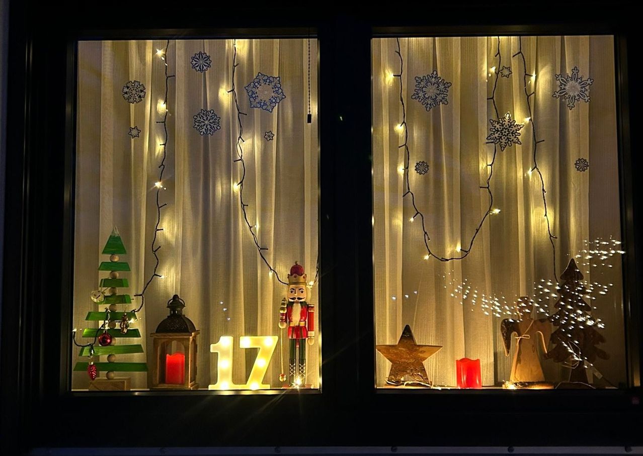Zwei Fensterdisplays mit weißen Vorhängen, Schneeflocken und Lichtern. Eines hat einen Weihnachtsbaum, eine Nussknacker und die Zahl '17'. Das andere hat einen Stern, einen Engel und eine Kerze.