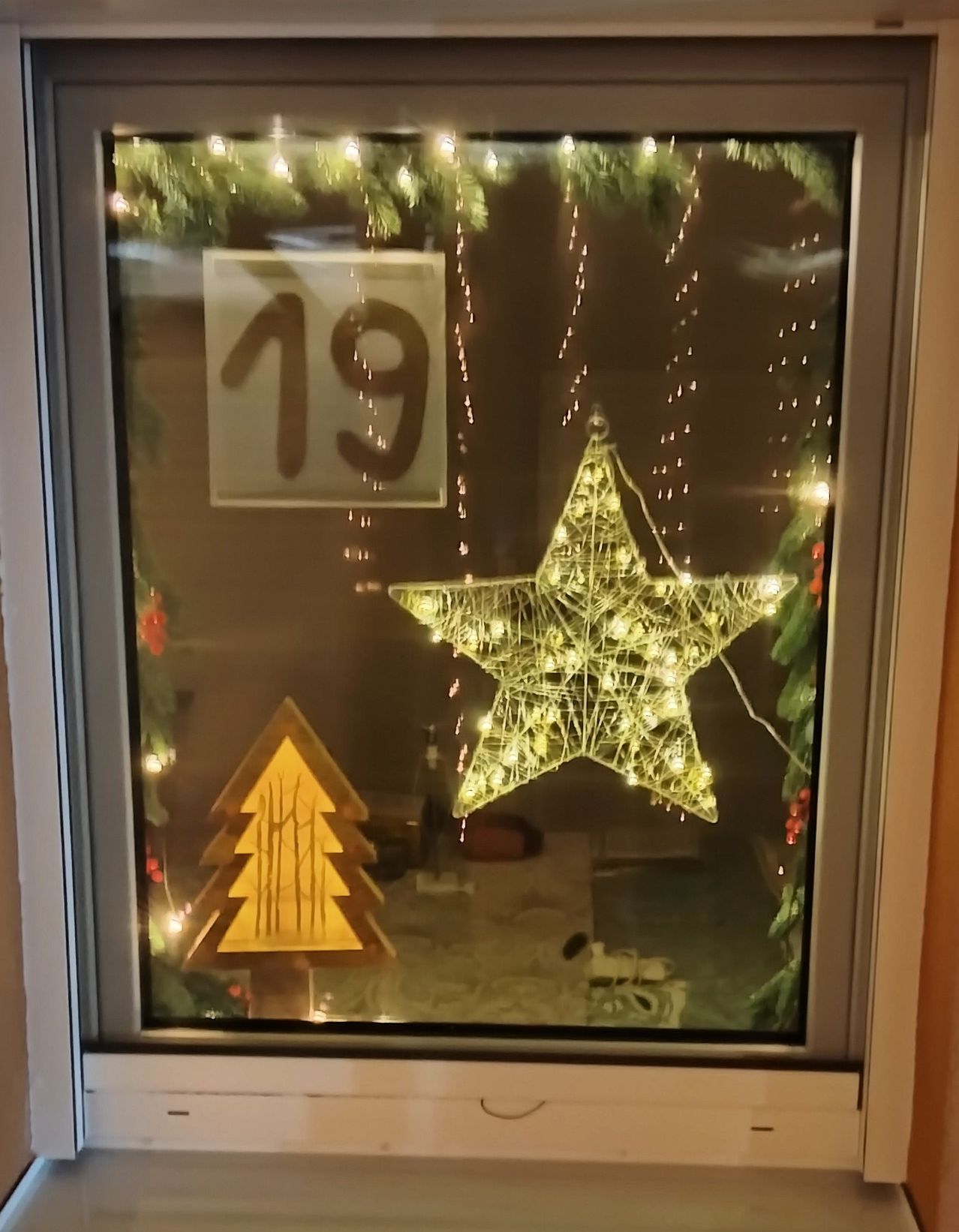 Ein Fenster zeigt eine Weihnachtsdekoration mit einem Stern aus Lichterketten und der Nummer 19. Ein Weihnachtsbaum und einige Weihnachtslichter sind ebenfalls sichtbar.