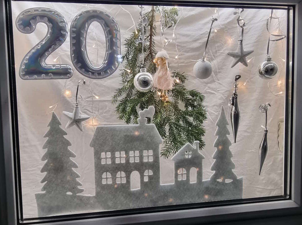 Eine Fensterdekoration zeigt eine festliche Szene mit einem Weihnachtsbaum, Ornamenten und der Nummer 20 darüber. Darunter befinden sich Häuser und Bäume aus lichtdurchlässigem Material.
