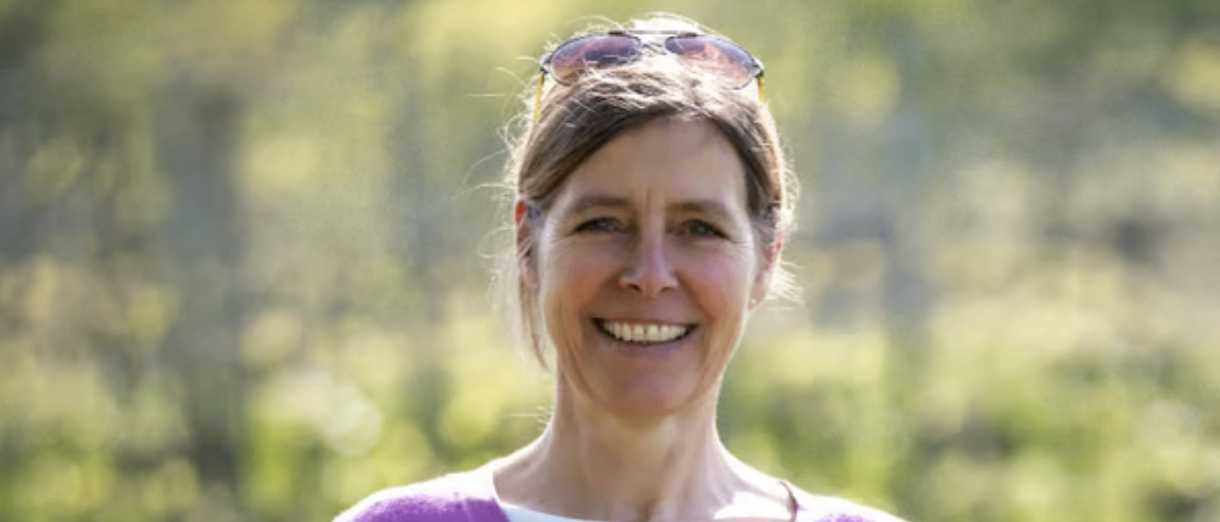 Eine lächelnde Frau mit Sonnenbrille auf dem Kopf steht draußen und trägt ein lila V-Neck-Shirt. Der Hintergrund ist unscharf mit grünem Laub.