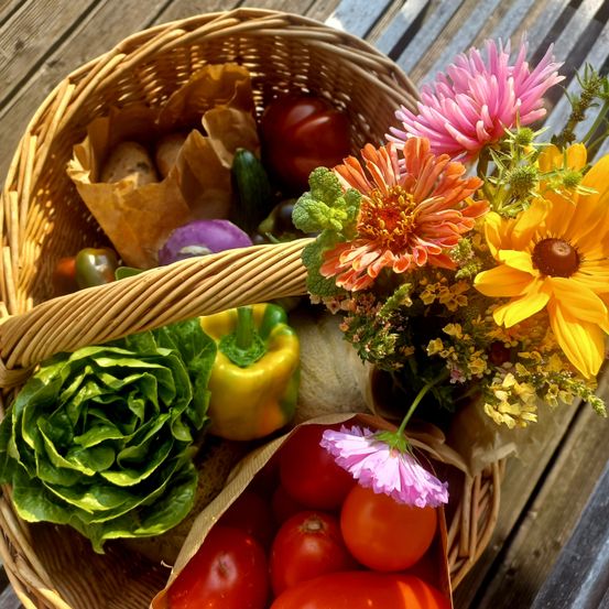 Ein Korb aus Weide gefüllt mit verschiedenen Gemüsesorten und Blumen. Tomaten, Paprika und Gurken sind im Inneren. Es gibt auch Rosen und Sonnenblumen. Der Korb steht auf einem Holztisch.