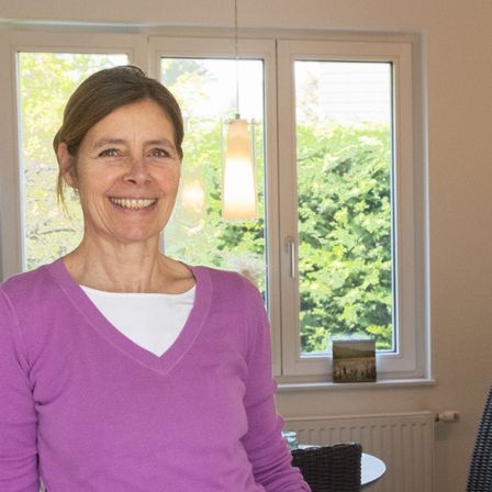 Eine lächelnde Frau in einem lila Shirt steht drinnen, mit großen Fenstern hinter ihr, die Grün draußen zeigen.
