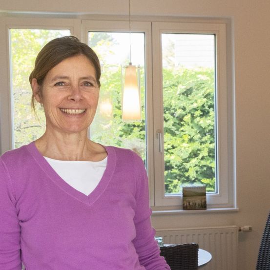 Eine lächelnde Frau in einem lila Shirt steht drinnen, mit großen Fenstern hinter ihr, die Grün draußen zeigen.